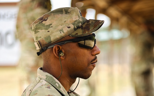 A U.S. Army soldier in Operational Camouflage Pattern (OCP) uniform is photographed in profile view at what appears to be a training range or outdoor military facility. The soldier is wearing a patrol cap, ballistic eyewear, and an in-ear communication device, and has a 'T14' identifier patch on their left sleeve. They are holding a small black pouch or device in their hands.