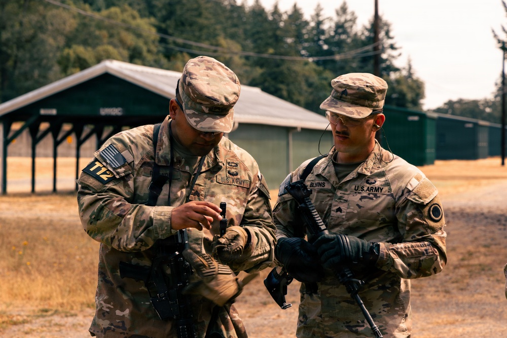 Two U.S. Army soldiers in OCP (Operational Camouflage Pattern) uniforms are standing outdoors at what appears to be a military training facility. The soldier on the left, wearing a unit patch 'T12', appears to be inspecting or adjusting equipment while the soldier on the right, identified by the name tape 'BURINS', observes. Both soldiers are armed and wearing tactical gloves.