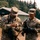 Two U.S. Army soldiers in OCP (Operational Camouflage Pattern) uniforms are standing outdoors at what appears to be a military training facility. The soldier on the left, wearing a unit patch 'T12', appears to be inspecting or adjusting equipment while the soldier on the right, identified by the name tape 'BURINS', observes. Both soldiers are armed and wearing tactical gloves.