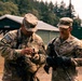 Two U.S. Army soldiers in OCP (Operational Camouflage Pattern) uniforms are standing outdoors at what appears to be a military training facility. The soldier on the left, wearing a unit patch 'T12', appears to be inspecting or adjusting equipment while the soldier on the right, identified by the name tape 'BURINS', observes. Both soldiers are armed and wearing tactical gloves.