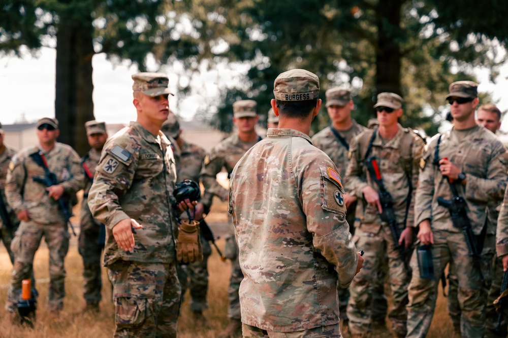 A group of U.S. Army soldiers in OCP (Operational Camouflage Pattern) uniforms are gathered outdoors in what appears to be a field training environment. A soldier facing the camera appears to be briefing or debriefing the group, while another soldier with the name tape 'BURGESS' stands with his back to the camera. The setting includes dry grass and trees, suggesting a training area or military installation.