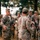 A group of U.S. Army soldiers in OCP (Operational Camouflage Pattern) uniforms are gathered outdoors in what appears to be a field training environment. A soldier facing the camera appears to be briefing or debriefing the group, while another soldier with the name tape 'BURGESS' stands with his back to the camera. The setting includes dry grass and trees, suggesting a training area or military installation.