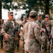 A group of U.S. Army soldiers in OCP (Operational Camouflage Pattern) uniforms are gathered outdoors in what appears to be a field training environment. A soldier facing the camera appears to be briefing or debriefing the group, while another soldier with the name tape 'BURGESS' stands with his back to the camera. The setting includes dry grass and trees, suggesting a training area or military installation.
