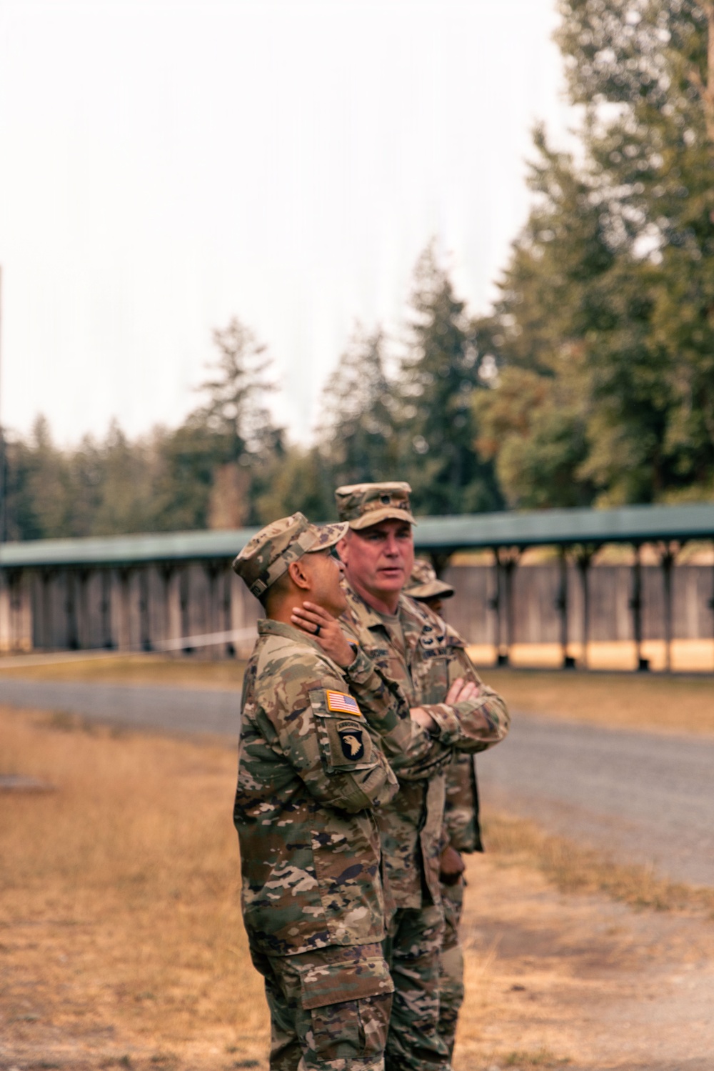 Two U.S. Army soldiers in OCP (Operational Camouflage Pattern) uniforms stand outdoors on what appears to be a military installation or training area. The foreground soldier displays a 101st Airborne Division patch and U.S. flag on the left sleeve, while both appear to be engaged in conversation or observation. A third soldier is partially visible in the background near a covered structure.
