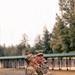 Two U.S. Army soldiers in OCP (Operational Camouflage Pattern) uniforms stand outdoors on what appears to be a military installation or training area. The foreground soldier displays a 101st Airborne Division patch and U.S. flag on the left sleeve, while both appear to be engaged in conversation or observation. A third soldier is partially visible in the background near a covered structure.