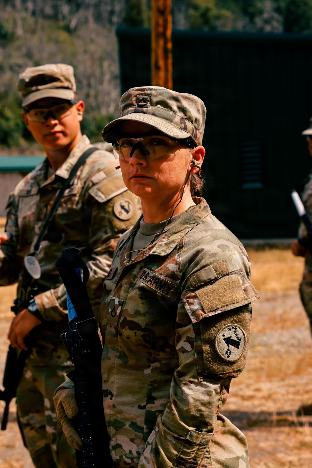 Two U.S. Army soldiers in Operational Camouflage Pattern (OCP) uniforms are standing at what appears to be a military training range. The foreground soldier is a female wearing protective eyewear and holding a rifle with a blue training barrel insert, indicating a live-fire or weapons qualification exercise. A second soldier is visible in the background, also in OCP uniform with unit patches visible.