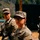 Two U.S. Army soldiers in Operational Camouflage Pattern (OCP) uniforms are standing at what appears to be a military training range. The foreground soldier is a female wearing protective eyewear and holding a rifle with a blue training barrel insert, indicating a live-fire or weapons qualification exercise. A second soldier is visible in the background, also in OCP uniform with unit patches visible.