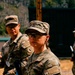 Two U.S. Army soldiers in Operational Camouflage Pattern (OCP) uniforms are standing at what appears to be a military training range. The foreground soldier is a female wearing protective eyewear and holding a rifle with a blue training barrel insert, indicating a live-fire or weapons qualification exercise. A second soldier is visible in the background, also in OCP uniform with unit patches visible.