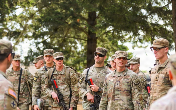 A group of U.S. Army soldiers in OCP (Operational Camouflage Pattern) uniforms are gathered outdoors in a field setting near trees, appearing to receive a briefing or instructions. Several soldiers are carrying rifles with red training/safety indicators attached, suggesting this is a training exercise. The soldiers are attentive and facing toward individuals on the left side of the frame who appear to be addressing the group.