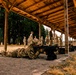 Military personnel are conducting a weapons qualification or marksmanship training exercise at an outdoor covered firing range. One soldier in the foreground is in the prone position aiming a rifle downrange, while other personnel stand at attention or observe along the firing line. The setting appears to be a standard military training installation with a wooden-roofed range structure.