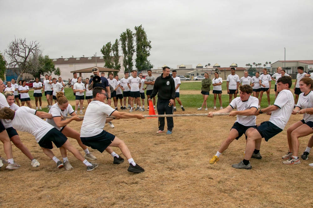 A group of young people in matching white t-shirts and dark shorts are engaged in a tug-of-war competition on a dry grass field. An adult supervisor in a dark jacket stands in the center observing the activity, while a large crowd of similarly dressed participants watches from the background. The setting appears to be a military or ROTC training event based on uniforms and organized structure.