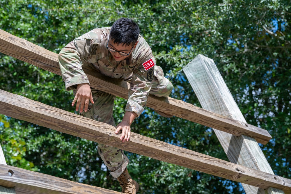A U.S. military service member wearing OCP (Operational Camouflage Pattern) uniform with a name tape reading 'TAO' and a red numbered bib '338' is navigating a wooden obstacle course. The individual is climbing or traversing horizontal wooden beams, suggesting participation in a physical fitness or training evaluation. The setting appears to be an outdoor military training facility surrounded by trees.