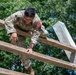 A U.S. military service member wearing OCP (Operational Camouflage Pattern) uniform with a name tape reading 'TAO' and a red numbered bib '338' is navigating a wooden obstacle course. The individual is climbing or traversing horizontal wooden beams, suggesting participation in a physical fitness or training evaluation. The setting appears to be an outdoor military training facility surrounded by trees.