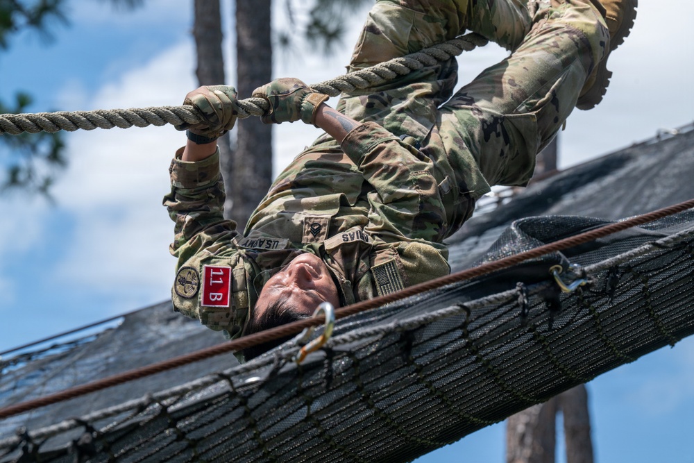 A U.S. Army soldier in Operational Camouflage Pattern (OCP) uniform is navigating an overhead rope obstacle on a military obstacle course. The soldier is hanging upside down beneath a cargo net structure, gripping a rope while traversing the course. Patches visible on the uniform include 'US ARMY,' a name tape reading 'RIAS,' a red '11B' infantry patch, and a U.S. flag patch.