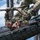 A U.S. Army soldier in Operational Camouflage Pattern (OCP) uniform is navigating an overhead rope obstacle on a military obstacle course. The soldier is hanging upside down beneath a cargo net structure, gripping a rope while traversing the course. Patches visible on the uniform include 'US ARMY,' a name tape reading 'RIAS,' a red '11B' infantry patch, and a U.S. flag patch.