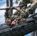 A U.S. Army soldier in Operational Camouflage Pattern (OCP) uniform is navigating an overhead rope obstacle on a military obstacle course. The soldier is hanging upside down beneath a cargo net structure, gripping a rope while traversing the course. Patches visible on the uniform include 'US ARMY,' a name tape reading 'RIAS,' a red '11B' infantry patch, and a U.S. flag patch.