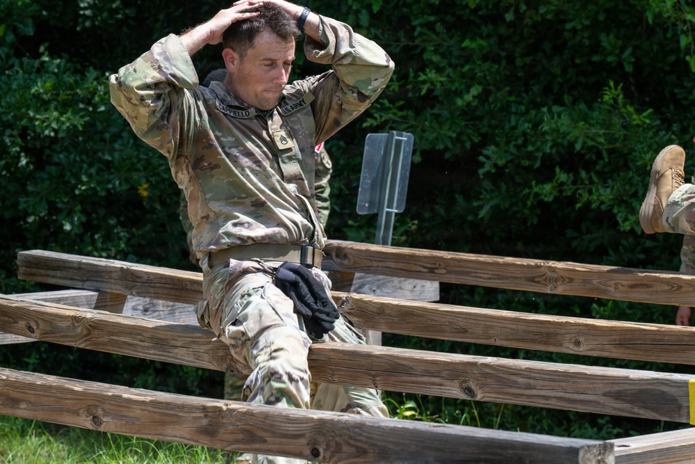 A U.S. Army soldier named Cappiello is navigating a wooden obstacle course, appearing to climb over or through horizontal wooden rails. The soldier is wearing full OCP (Operational Camouflage Pattern) uniform with hands placed behind his head, suggesting a specific movement technique or exercise requirement. A second soldier is partially visible in the background to the right.