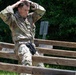 A U.S. Army soldier named Cappiello is navigating a wooden obstacle course, appearing to climb over or through horizontal wooden rails. The soldier is wearing full OCP (Operational Camouflage Pattern) uniform with hands placed behind his head, suggesting a specific movement technique or exercise requirement. A second soldier is partially visible in the background to the right.