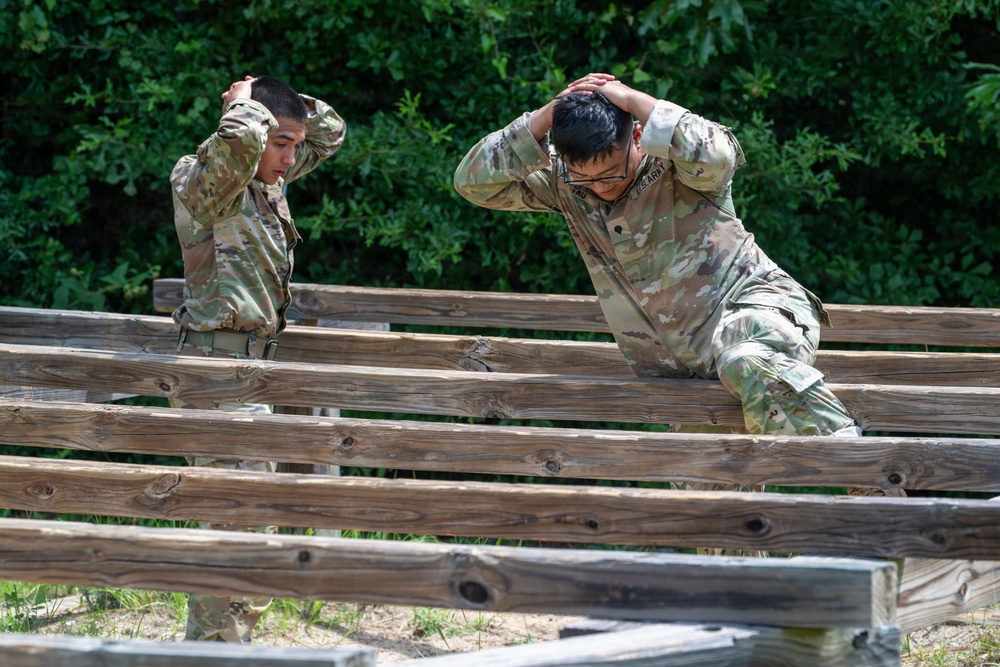 Two U.S. Army soldiers in MultiCam/OCP camouflage uniforms are navigating a wooden obstacle course, both with hands placed on the backs of their heads. The soldier on the right is wearing glasses and has a visible 'U.S. ARMY' tape on their uniform. They appear to be conducting physical training or an obstacle course assessment in an outdoor wooded environment.
