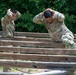 Two U.S. Army soldiers in MultiCam/OCP camouflage uniforms are navigating a wooden obstacle course, both with hands placed on the backs of their heads. The soldier on the right is wearing glasses and has a visible 'U.S. ARMY' tape on their uniform. They appear to be conducting physical training or an obstacle course assessment in an outdoor wooded environment.