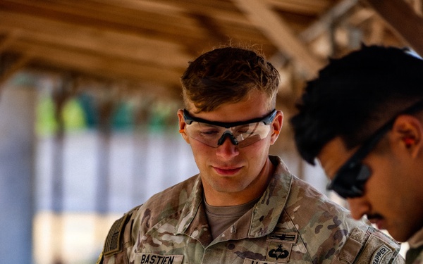 A U.S. Army Staff Sergeant named Bastien, wearing OCP (Operational Camouflage Pattern) uniform and ballistic eyewear, stands under a wooden-roofed structure while another soldier appears to be checking or adjusting something at his waist. The soldier displays a Staff Sergeant rank insignia on his chest and what appears to be an Engineer or similar badge above the U.S. Army tape. A second soldier to the right wears a 'SAPPER' tab on their shoulder sleeve.