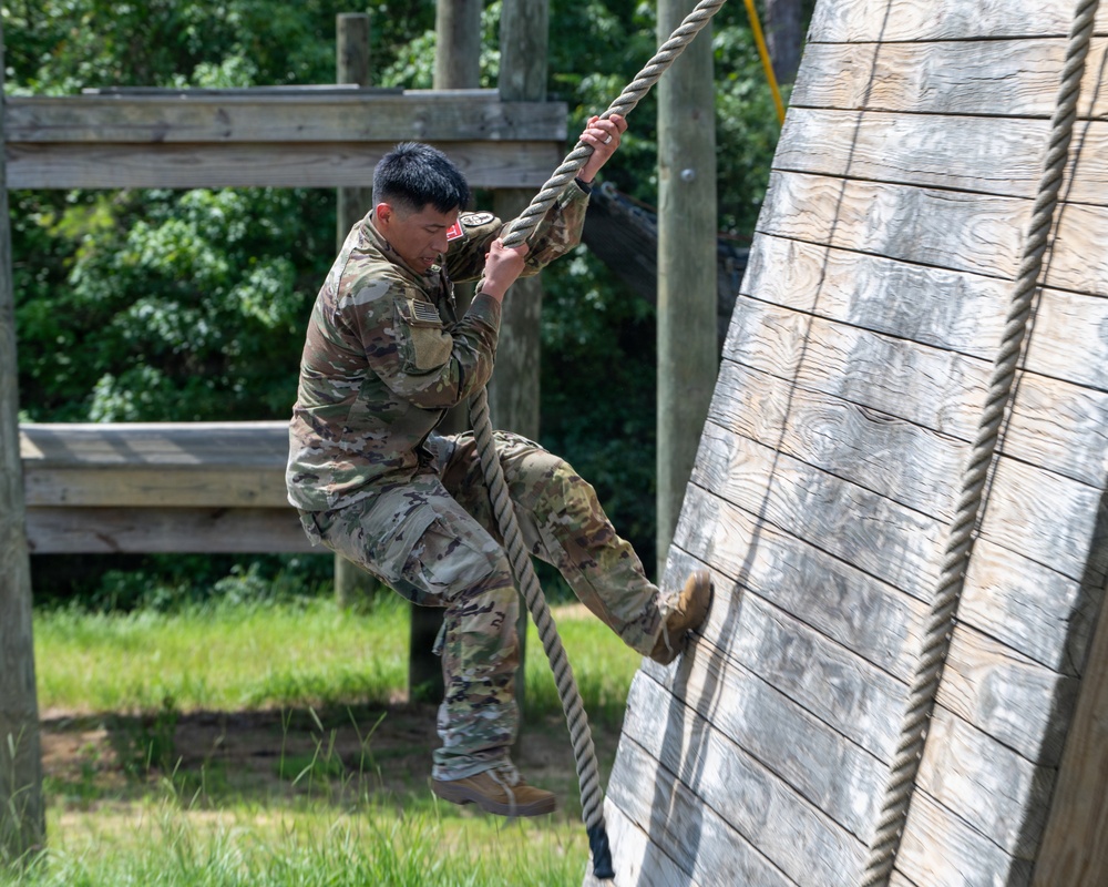 A U.S. Army soldier in OCP (Operational Camouflage Pattern) uniform is climbing a rope on a military obstacle course. The soldier is using both hands and feet to ascend the rope alongside an angled wooden wall structure. The setting appears to be an outdoor training facility with wooden obstacles and lush green vegetation in the background.