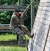 A U.S. Army soldier in OCP (Operational Camouflage Pattern) uniform is climbing a rope on a military obstacle course. The soldier is using both hands and feet to ascend the rope alongside an angled wooden wall structure. The setting appears to be an outdoor training facility with wooden obstacles and lush green vegetation in the background.