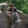 Three military personnel in OCP (Operational Camouflage Pattern) uniforms are navigating a wooden wall obstacle, likely part of a military training course. Two soldiers at the top of the wall are pulling up a third soldier from below, demonstrating teamwork and physical fitness training. The setting appears to be an outdoor obstacle course surrounded by green foliage.