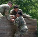 Three military personnel in OCP (Operational Camouflage Pattern) uniforms are navigating a wooden wall obstacle, likely part of a military training course. Two soldiers at the top of the wall are pulling up a third soldier from below, demonstrating teamwork and physical fitness training. The setting appears to be an outdoor obstacle course surrounded by green foliage.
