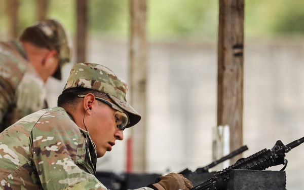 A soldier in MultiCam/OCP camouflage uniform is in a prone firing position at what appears to be a military shooting range. The soldier is aiming a rifle, likely an M4 or M16 variant, over a concrete block barrier. Additional soldiers in similar uniforms are visible in the blurred background, suggesting a group training exercise.
