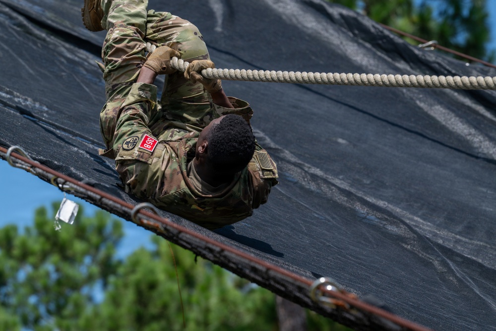 A soldier in US Army OCP (Operational Camouflage Pattern) uniform is climbing a steep inclined obstacle using a rope, lying on their back against a black tarp-covered surface. The soldier appears to be navigating a military obstacle course, gripping a thick rope for assistance. Pine trees and a blue sky are visible in the background, suggesting an outdoor training environment.