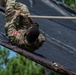 A soldier in US Army OCP (Operational Camouflage Pattern) uniform is climbing a steep inclined obstacle using a rope, lying on their back against a black tarp-covered surface. The soldier appears to be navigating a military obstacle course, gripping a thick rope for assistance. Pine trees and a blue sky are visible in the background, suggesting an outdoor training environment.