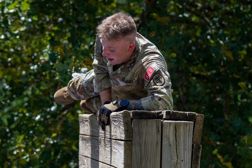 A U.S. Army soldier in MultiCam camouflage uniform is navigating an obstacle course, climbing over a large wooden wall or barrier. The soldier is wearing tactical gloves, a watch, and has a red patch with '33B' visible on the sleeve along with a unit insignia. The image was taken outdoors with green foliage visible in the background.