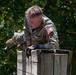 A U.S. Army soldier in MultiCam camouflage uniform is navigating an obstacle course, climbing over a large wooden wall or barrier. The soldier is wearing tactical gloves, a watch, and has a red patch with '33B' visible on the sleeve along with a unit insignia. The image was taken outdoors with green foliage visible in the background.