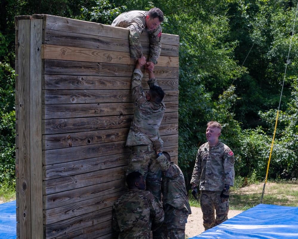 Military personnel in camouflage uniforms are completing a team obstacle course challenge involving scaling a tall wooden wall. One soldier has reached the top and is pulling up a second soldier by the hand, while a third soldier provides a boost from below. A fourth soldier stands nearby observing or waiting their turn.
