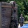 Military personnel in camouflage uniforms are completing a team obstacle course challenge involving scaling a tall wooden wall. One soldier has reached the top and is pulling up a second soldier by the hand, while a third soldier provides a boost from below. A fourth soldier stands nearby observing or waiting their turn.