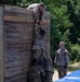 Military personnel in camouflage uniforms are completing a team obstacle course challenge involving scaling a tall wooden wall. One soldier has reached the top and is pulling up a second soldier by the hand, while a third soldier provides a boost from below. A fourth soldier stands nearby observing or waiting their turn.
