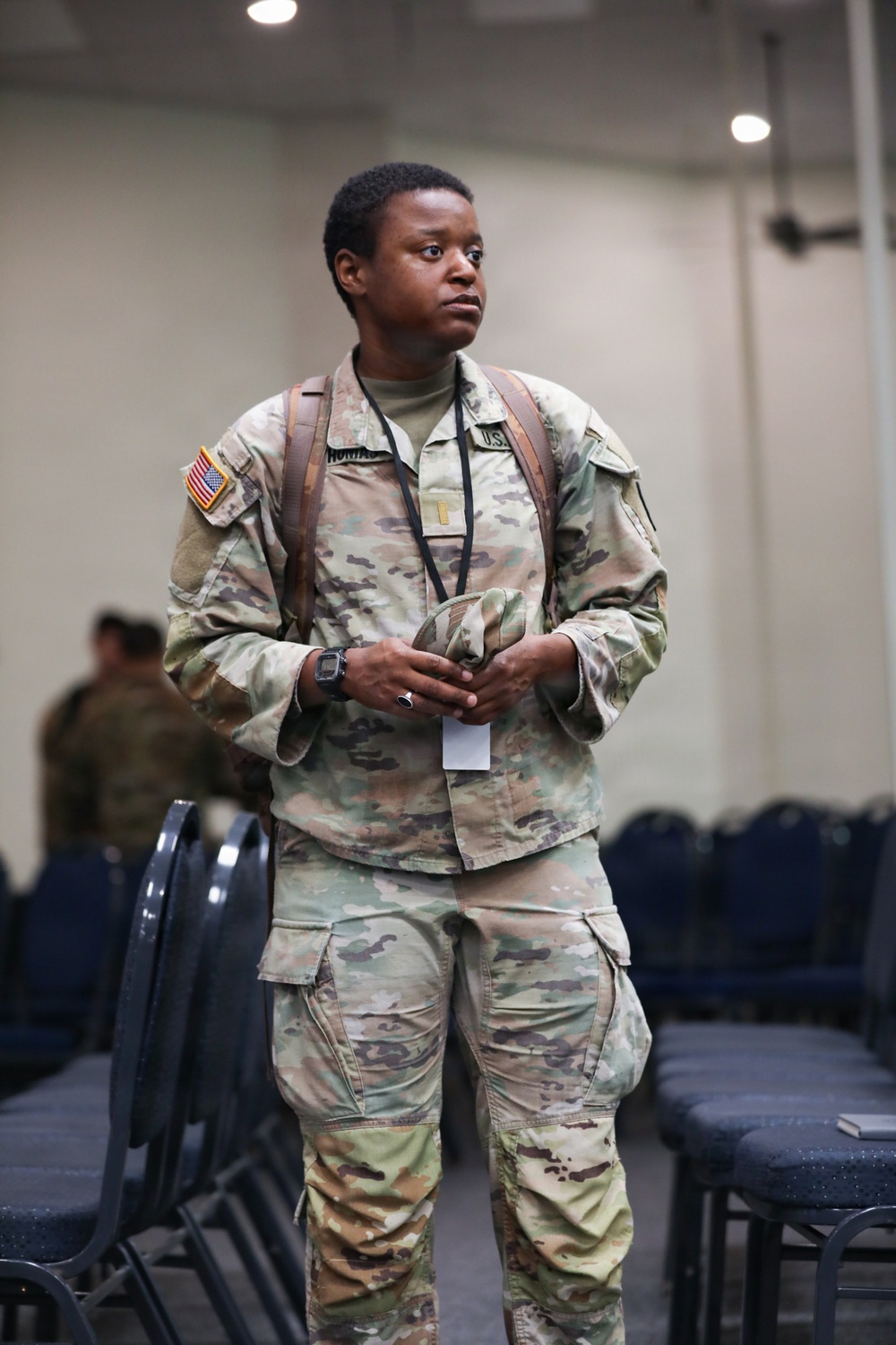 A U.S. Army soldier in Operational Camouflage Pattern (OCP) uniform stands in what appears to be a large indoor briefing or assembly hall. The soldier is wearing a lanyard with a badge, carrying a backpack, and holding a patrol cap while looking to the side. The setting contains rows of chairs, suggesting a formal gathering or event.