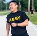 A person wearing a black 'ARMY' t-shirt is running and laughing with visible joy and exertion. The setting appears to be a military base or installation road, with other individuals visible in the background. The image captures an energetic, positive moment during what appears to be a physical training exercise.