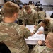 A group of U.S. Army soldiers in MultiCam/OCP camouflage uniforms are engaged in a classroom training exercise. They are gathered around a table reviewing a large paper worksheet related to medical evacuation (MEDEVAC) procedures, including fields for patient pickup location, radio frequency, and number of patients. The setting appears to be a military training facility with multiple small groups working simultaneously.