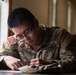 A U.S. Army soldier named Teague is seated at a desk writing or filling out paperwork. The soldier is wearing the standard Army Combat Uniform (ACU) in OCP (Operational Camouflage Pattern) with a U.S. flag patch on the left shoulder. The image is a close-up, well-lit photograph taken near a window.