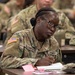 A U.S. Army soldier named Solomon is seated at a desk in what appears to be a classroom or briefing setting, taking notes or completing paperwork. She is wearing the standard Army Combat Uniform (ACU) in OCP (Operational Camouflage Pattern) with a U.S. flag patch on the left shoulder. Multiple other soldiers in identical uniforms are visible in the background, suggesting a group training or instruction session.