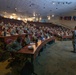 A military briefing or lecture is taking place in a tiered auditorium-style classroom, with a large group of uniformed soldiers seated at curved wooden desks facing an instructor standing at a podium. The attendees are wearing U.S. Army OCP (Operational Camouflage Pattern) uniforms. Multiple military unit flags are displayed along the right wall, indicating an official military educational or training facility.