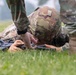 A soldier in MultiCam/OCP camouflage uniform is lying prone on grass, closely inspecting or working on a rifle. A second individual in matching camouflage is standing nearby, appearing to observe or instruct. The setting appears to be an outdoor training area with green grass and a blurred background.