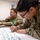 2026 Paralegal Warrior Two U.S. Army soldiers in OCP (Operational Camouflage Pattern) uniforms are engaged in a training or planning exercise. A female soldier in the foreground is writing on a large white paper/poster with a green marker, while a male soldier sits in the background. The setting appears to be an indoor classroom or conference room environment.