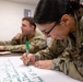 Two U.S. Army soldiers in OCP (Operational Camouflage Pattern) uniforms are engaged in a training or planning exercise. A female soldier in the foreground is writing on a large white paper/poster with a green marker, while a male soldier sits in the background. The setting appears to be an indoor classroom or conference room environment.
