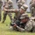 2026 Paralegal Warrior A U.S. Army instructor is coaching a soldier in the prone firing position on a grass field. The instructor, wearing glasses and identified by a name tape reading 'WU', is gesturing with an outstretched hand, likely indicating a target direction or correcting aim. Additional soldiers in OCP (Operational Camouflage Pattern) uniforms are visible in the blurred background observing the training.