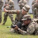 A U.S. Army instructor is coaching a soldier in the prone firing position on a grass field. The instructor, wearing glasses and identified by a name tape reading 'WU', is gesturing with an outstretched hand, likely indicating a target direction or correcting aim. Additional soldiers in OCP (Operational Camouflage Pattern) uniforms are visible in the blurred background observing the training.