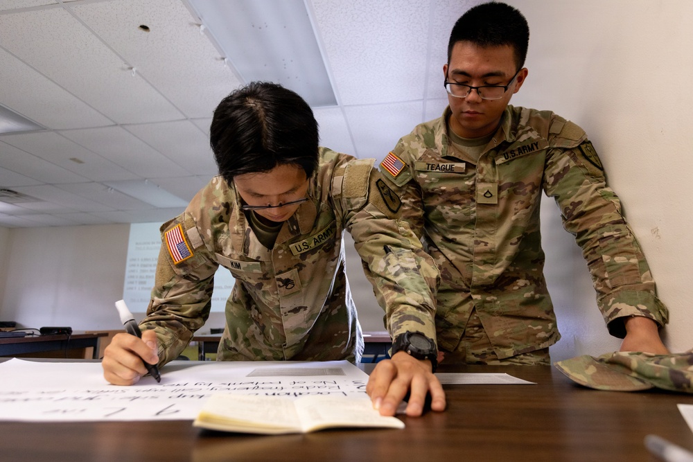 Two U.S. Army soldiers in OCP (Operational Camouflage Pattern) uniforms are working together at a table in what appears to be a classroom or training environment. The soldier on the left, identified by name tape as 'KIM,' is writing on a large sheet of paper, while the soldier on the right, identified as 'TEAGUE,' is leaning over and referencing notes. Both wear glasses and American flag patches on their left shoulders.