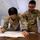 2026 Paralegal Warrior Two U.S. Army soldiers in OCP (Operational Camouflage Pattern) uniforms are working together at a table in what appears to be a classroom or training environment. The soldier on the left, identified by name tape as 'KIM,' is writing on a large sheet of paper, while the soldier on the right, identified as 'TEAGUE,' is leaning over and referencing notes. Both wear glasses and American flag patches on their left shoulders.